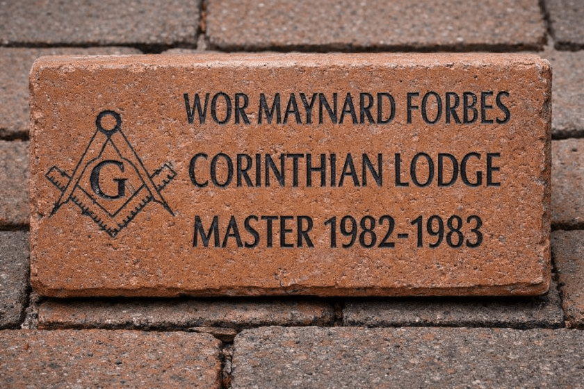 Engraved brick reads “WOR MAYNARD FORBES / CORINTHIAN LODGE / MASTER 1982–1983” with a square-and-compass symbol, set in a brick walkway.